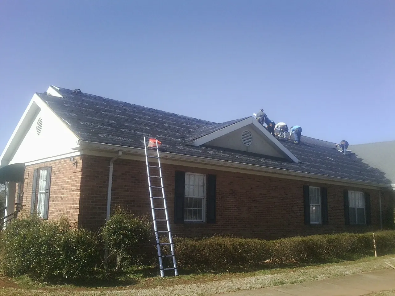 Crew tearing shingles off a brick ranch roof with a ladder set — Emergency Roof Repair work by our crew in Baldwin Place