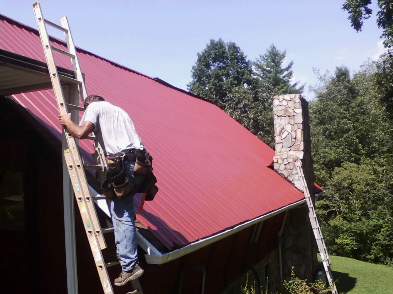 Roofer on a ladder beside a red metal roof and stone chimney — Soffit & Fascia Repair by Roofing Brothers in Baldwin Place